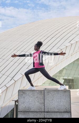 Seitenansicht einer fokussierten afroamerikanischen Sportlerin, die beim Training in der Stadt Ausfallschritte mit erhobenen Armen macht, während sie die Beine dehnt Stockfoto