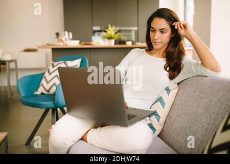 Junge Freiberuflerin im lässigen Outfit sitzt auf Sofa und Mit einem Laptop arbeiten, während Sie von zu Hause aus im Wohnzimmer arbeiten Stockfoto