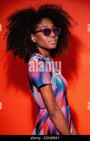 Moderne kubanische Frau mit Afro-Haaren trägt ein helles Kleid und eine Sonnenbrille im roten Studiohintergrund Stockfoto