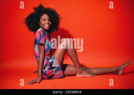 Moderne kubanische Frau mit Afro-Haaren in einem hellen Kleid und lächelnd vor rotem Studiohintergrund Stockfoto