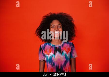 Moderne kubanische Frau mit Afro-Haaren in einem hellen Kleid und lächelnd vor rotem Studiohintergrund Stockfoto