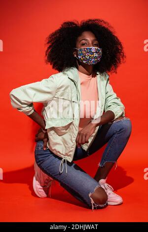 Junge ethnische Frau mit Afro-Frisur, die Stoffmaske trägt und auf rotem Hintergrund emotionslos auf die Kamera schaut Stockfoto