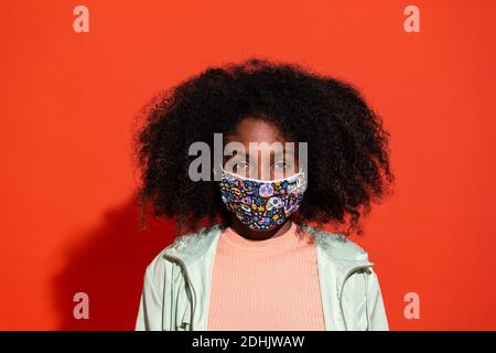 Junge ethnische Frau mit Afro-Frisur, die Stoffmaske trägt und auf rotem Hintergrund emotionslos auf die Kamera schaut Stockfoto