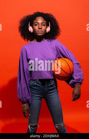 Moderne, unemotionale schwarze Frau mit Afro-Frisur, die rosa Kopfhörer trägt und orangen Basketball hält und Musik auf rotem Hintergrund hört Stockfoto