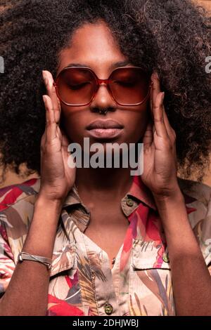 Selbstbewusste kubanische Frau mit Afro-Frisur trägt eine Sonnenbrille mit einem floralen Hemd, das die Augen schließt Stockfoto