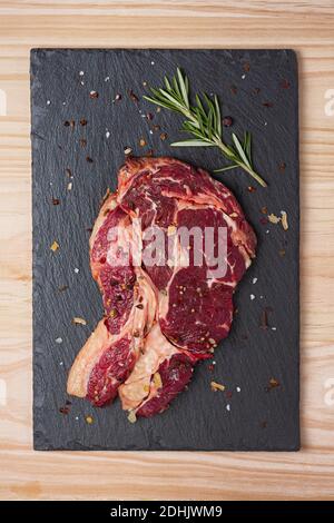 Blick von oben auf das frische, ungekochte Rindersteak mit Rib Eye Rosmarinzweig und getrocknete Gewürze auf schwarzem Schieferbrett Stockfoto