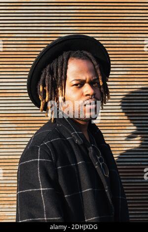 Moderne Erwachsene schwarzen Mann mit Dreadlocks tragen Hut und stilvoll Mantel Blick auf die Kamera gegen die Straßenwand im Sonnenlicht Stockfoto