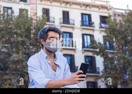 Ernst männlich in stilvoller Kleidung und Schutzmaske in der Stadt Und SMS-Nachrichten auf Handy während Coronavirus Ausbruch Stockfoto