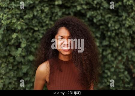 Optimistische afroamerikanische junge Frau mit lockigen Haaren und charmant Lächeln Sie und schauen Sie auf die Kamera, während Sie im Park gegen das Grün stehen Anlagen Stockfoto