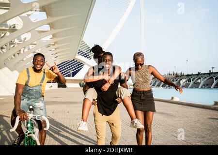 Fröhliche Gesellschaft afroamerikanischer Freunde, die Longboard und Fahrrad fahren In der Nähe Huckepack Paar, während Spaß und Unterhaltung in der Stadt Im Sommer Stockfoto