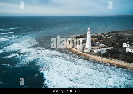 Von oben Drohne Blick auf den weißen Leuchtturm Turm und klein Siedlung an der felsigen Küste von schäumenden Meereswellen gewaschen Stockfoto