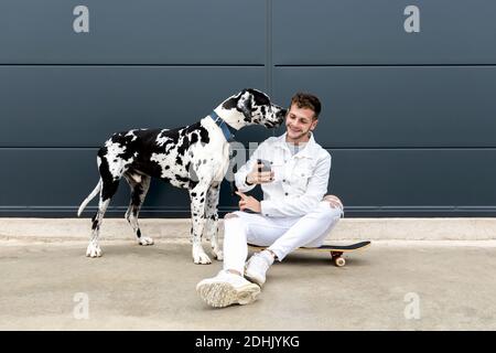 Fröhlicher junger Rüde kuschelt Dogge und nimmt sich selbst. Porträt auf dem Smartphone, während Sie zusammen in der Stadt sitzen Stockfoto