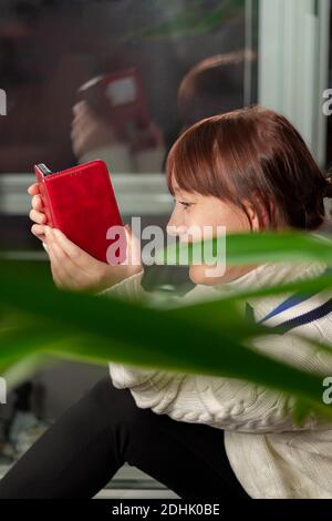 Ein junges hübsches Mädchen in einem weißen Pullover mit einem Smartphone in den Händen kommuniziert in einem Messenger zu Hause Am Fenster Stockfoto