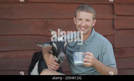 Der Bauer trinkt Ziegenmilch aus einem Becher und umarmt seine geliebte Ziege. Stockfoto
