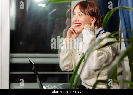 Junge hübsche Mädchen in Kopfhörer hört Musik mit Vergnügen Beim Sitzen auf der Fensterbank zu Hause am Fenster Stockfoto