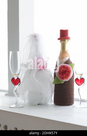 Zwei Hochzeitsflaschen Champagner in den Kostümen der Braut und des Bräutigams und zwei Gläser mit Herzen befinden sich am Fenster auf der Fensterbank. Mi Stockfoto