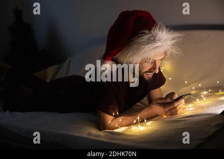 Hübscher junger Mann, der auf der Couch liegt und einen Weihnachtsmütze trägt, sein Telefon benutzt und in einem dunklen weihnachtlich dekorierten Zimmer lächelt. Es ist angenehm, Zeit zu verbringen Stockfoto