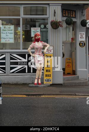 Weibliche Schaufensterpuppe in rot und weiß Kellnerin Uniform gekleidet tragen Ein Schal als Gesichtsmask außerhalb eines Chipshops Stockfoto