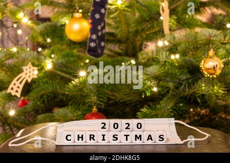 Symbol für die Weihnachtszeit während der Corona-Pandemie. Würfel bilden den Ausdruck "Weihnachten 2020" auf einer Gesichtsmaske vor einem Weihnachtsbaum Stockfoto