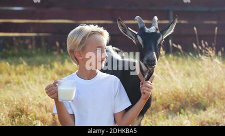 Ein Junge trinkt Ziegenmilch aus einem Becher neben seiner Ziege. Stockfoto