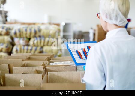 Junge weibliche fokussierte Arbeiterin in sterilen Tüchern prüft die Charts Aus ihrem Ordner, während sie in der Nähe der Kisten in der Fabrik stand Lagerbereich und Zählung Th Stockfoto