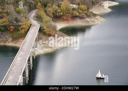 Ein kleines Boot segelt auf dem Turanosee in der Nähe der Brücke, die zum alten Dorf Castel di Tora führt. Castel di Tora, Rieti, Latium, Italien. Stockfoto