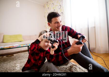 Junge fröhlich aufgeregt Vater und Sohn in der gleichen roten shirt spielen Konsolenspiele mit Gamepads, während sich an jedem lehnt Andere in einem hellen Wohn ro Stockfoto