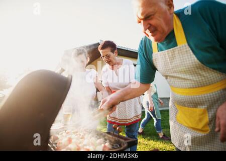 Großmutter bereitet andere Gerichte mit ihrer Enkelin zu, während vorsichtiger Großvater während des Grills Fleisch vorsichtig dreht. Stockfoto