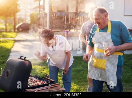 Ältere Frau inspiziert sorgfältig, sollte sie Fleisch vom Grill nehmen, während ihr Mann neben ihr steht und lächelt. Stockfoto