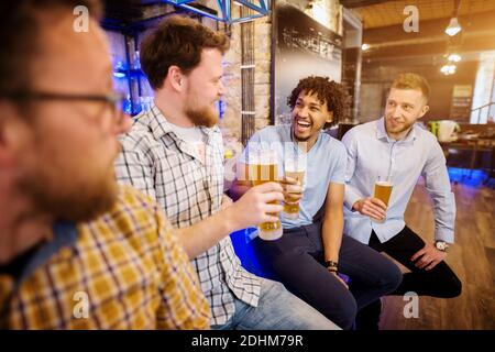 Lachende multikulturelle Freunde sitzen auf dem Barstuhl und lauschen der Freundschaftsgeschichte mit Bier vom Fass in den Händen. Stockfoto