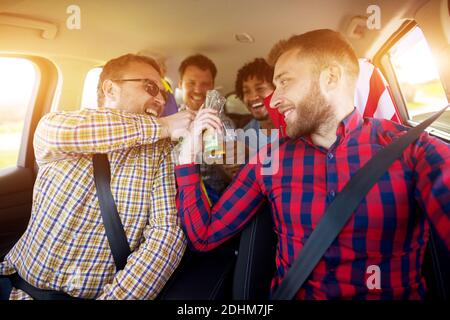 Junge fröhliche multikulturelle Freunde, die mit einem Bier im Auto auf der Fahrt klirren. Stockfoto