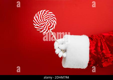 Weihnachtsmann Hand mit bunten Lollipop auf rotem Hintergrund, Holzstab, rote und weiße Spirale, Kinder Süßigkeiten, Weihnachtskonzept Stockfoto