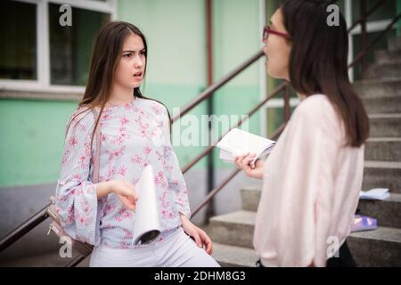 Zwei verärgerte High School Mädchen, die Missverständnisse hatten, während sie vor der Schule mit Notizen in den Händen standen. Stockfoto