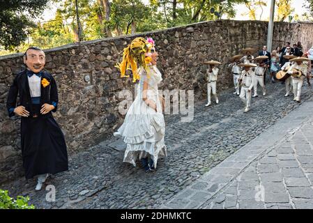 Eine traditionelle mexikanische Mariachi-Band spielt als riesige Puppen namens Mojigangas Tanz während einer Hochzeitsfeier Paraden durch die Straßen San Miguel de Allende, Guanajuato, Mexiko. Stockfoto