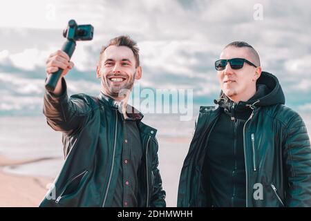 Zwei gutaussehende lächelnde Freunde machen Selfie mit Action-Kamera mit Gimbal Stabilisator am Strand. Jugendliche Männer in schwarzen Kleidern Spaß haben, indem Sie Fotos Stockfoto