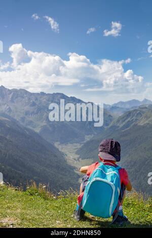 Abenteuerlicher und unerschrockener kleiner Junge mit blauer Mütze und Rucksack, der auf dem Rücken hockt, beobachtet von der Spitze eines Berges das Tal eines Berges Stockfoto