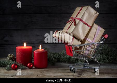 Kleiner Einkaufswagen voll mit Geschenken, zwei roten Kerzen, Weihnachtsdekoration und Tannenzweigen auf einem rustikalen Holztisch, dunkler Hintergrund mit Kopie s Stockfoto