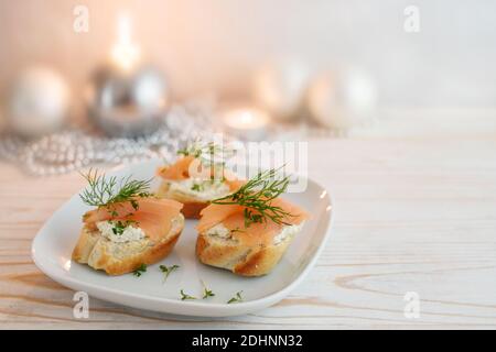 Festliche Canapés aus Baguette-Brot mit geräuchertem Lachs, Frischkäse und Dill garnieren auf einem Teller, weiß bemalter Holztisch mit Kerzen und Christma Stockfoto