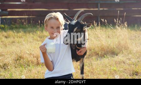 Ein Junge trinkt Ziegenmilch aus einem Becher neben seiner Ziege. Stockfoto