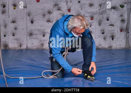 Professioneller älterer Mann trägt eine schützende Gesichtsmaske, die seine Kletterschuhe bindet, um sich auf eine künstliche Kletterwand vorzubereiten. Extreme Spor Stockfoto