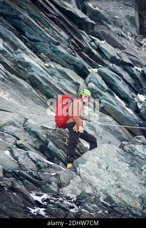 Glücklicher Mann in Wandersonnenbrille, der auf extrem senkrechten Felsen steht und Seil hält. Männlicher Reisender mit Rucksack und Blick auf die Kamera und lächelnd. Konzept von Bergsteigen, Alpinismus und Klettern. Stockfoto