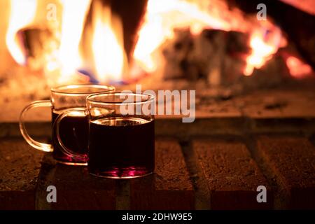 Zwei Tassen Wein oder Glühwein am Feuer. Die Tassen stehen an einem Ziegelkamin mit einem Feuer im Hintergrund, und es gibt Kopierplatz in der Stockfoto