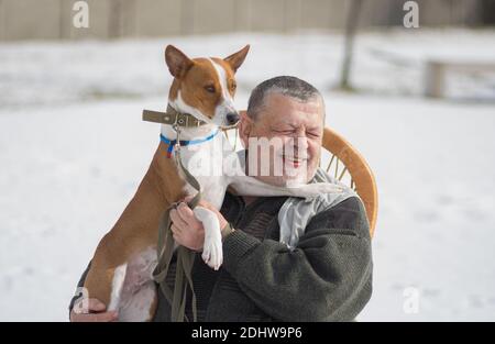Outdoor-Porträt des lächelnden kaukasischen älteren Mann, der seinen niedlichen basenji-Hund auf die Hände nimmt, während er in einem Korbstuhl in der Wintersaison sitzt. Stockfoto
