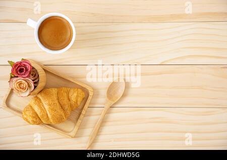 Ein Stück Buttercroissant auf einem Holzteller und eine Tasse Kaffee in einem weißen Keramikbecher. Holzhintergrund. Stockfoto