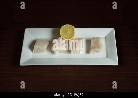 Vier Stücke frisches, rohes Seehechtfilet und eine Zitrone auf einem weißen Teller auf einem Holztisch in zwei Hälften geschnitten. Zutaten und Rezepte zum Kochen. Stockfoto