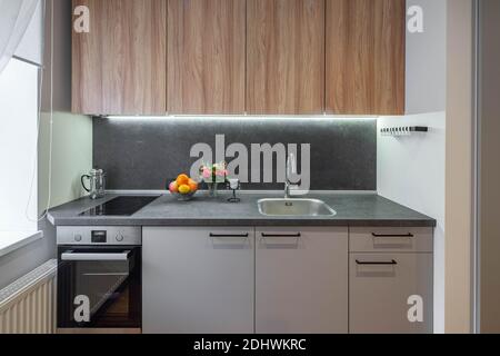 Modernes Interieur der kleinen Küche in der neuen Wohnung. Graue Küche mit Waschbecken und Backofen. Holzschränke. Stockfoto