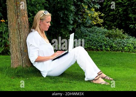 Eine junge Frau sitzt mit einem Laptop Computer im Garten und surft im Internet. WLAN im Park. Stockfoto