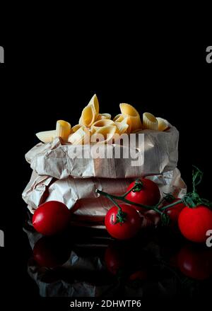 Frische Penne rigate Makkaroni Pasta in einem Papierbeutel und Kirschtomaten auf schwarzem Hintergrund Stockfoto