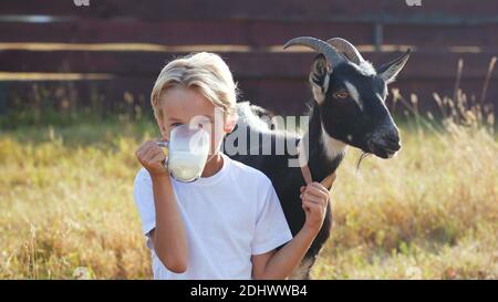 Ein Junge trinkt Ziegenmilch aus einem Becher neben seiner Ziege. Stockfoto