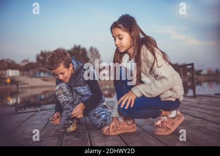 Zwei kleine, niedliche kleine Freunde, Junge und Mädchen, die abends auf einem See angeln. Kleiner Junge, der seinem Freund einen gefangenen Fisch zeigt. Kinder spielen. Friendshi Stockfoto